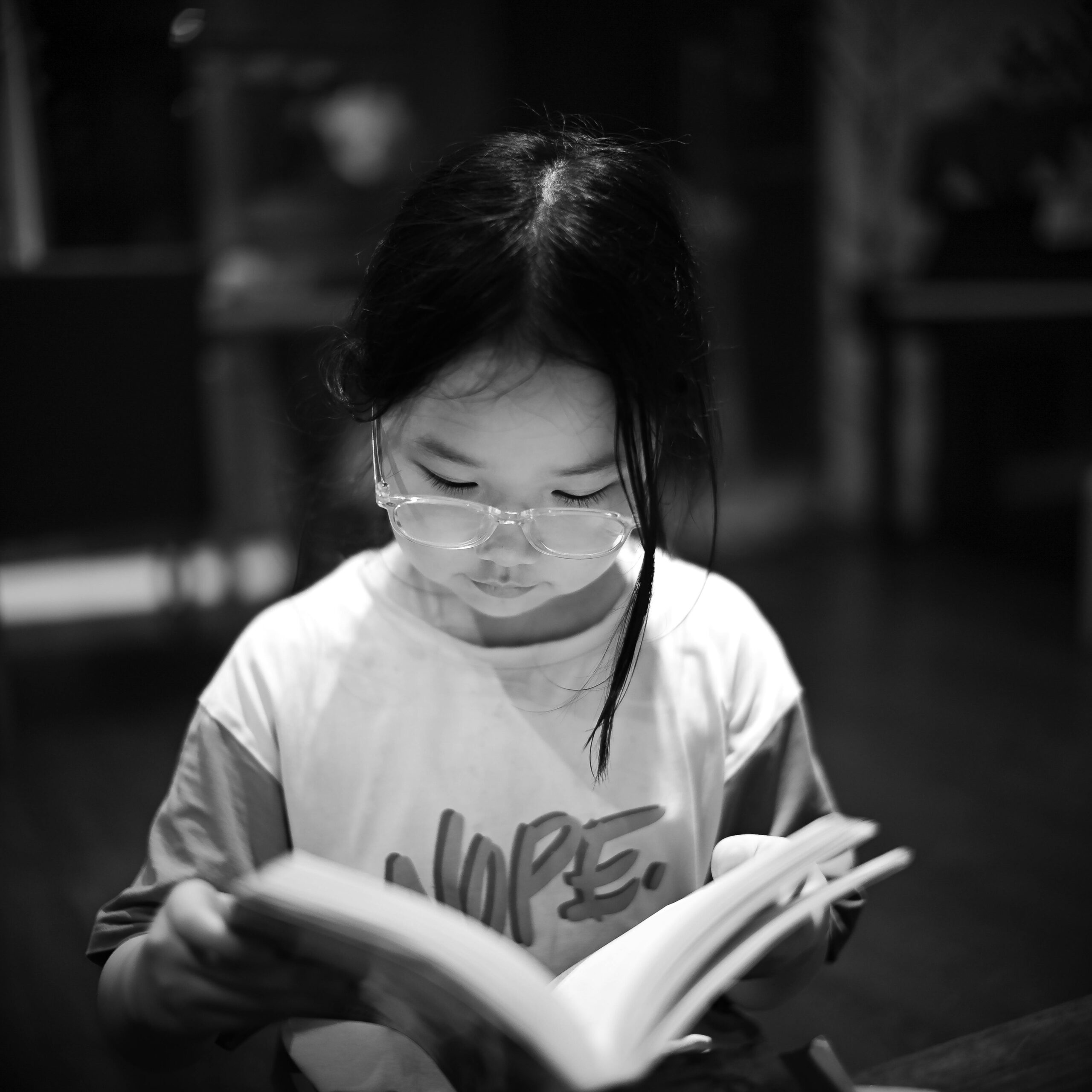 A young girl reading a book indoors, captured in black and white.